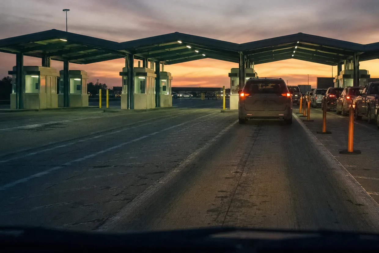 A land border crossing booth at dusk photographed from a driver's queue, fluorescent light spilling onto the empty lanes