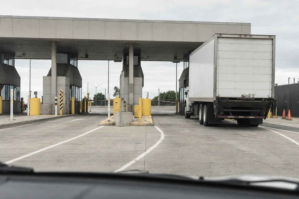A traveller's-eye view of a busy Canada-US border crossing plaza under flat grey skies, inspection booths visible ahead.