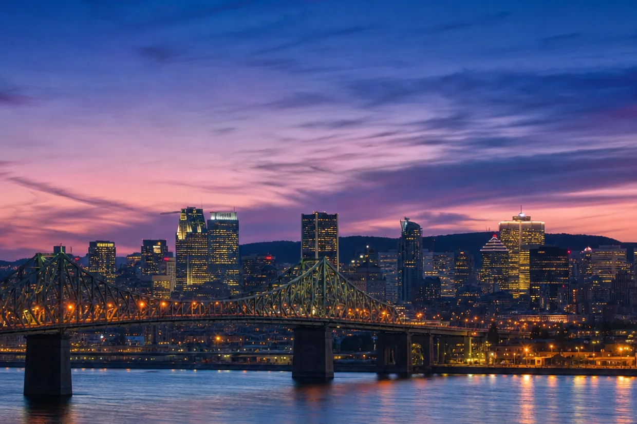 The Montreal skyline rises above the St. Lawrence River at dusk, the illuminated Jacques Cartier Bridge arching toward the
